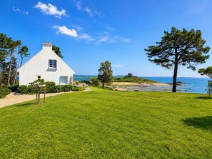 A white house with a small window and a door, surrounded by a well-maintained lawn and trees, with a clear view of the sea.