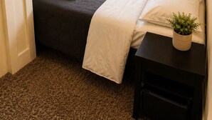 Une petite chambre à coucher, bien rangée, avec un lit, une table de chevet ornée d’une plante et une fenêtre avec des rideaux.