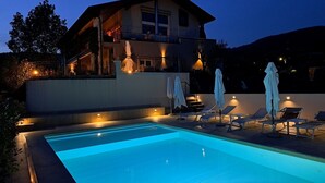 Una piscina con lettini e ombrelloni, circondata da un edificio con un balcone e illuminazione esterna.