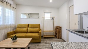 Un espacio habitable compacto con sofá, una mesita con planta, kitchenette con lavabo y ventana con cortinas translúcidas.