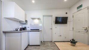 Cocina compacta con armarios blancos, microondas y cafetera. Hay una mesa de madera con una planta, un televisor montado en la pared y una puerta con la leyenda 'SALIDA DE EMERGENCIA'.