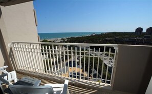 Un balcon avec des balustrades blanches, deux fauteuils de jardin blancs et une vue sur la plage et le paysage urbain.