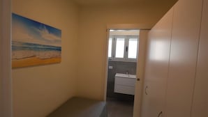 Een smalle gang met een schilderij van een strand aan de linker muur, die leidt naar een badkamer met een wastafel en kastjes.
