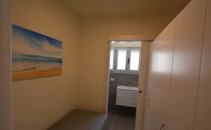 Un couloir étroit, avec une peinture représentant une plage sur le mur de gauche, mène à une salle de bain équipée d’un lavabo et de meubles de rangement.