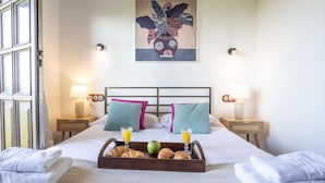 Una habitación de hotel con cama, almohadas, una bandeja con croissants y jugo, y un cuadro en la pared.