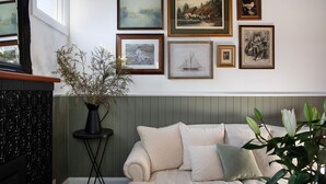A room with a beige sofa, a black vase with branches, and a wall decorated with framed pictures.