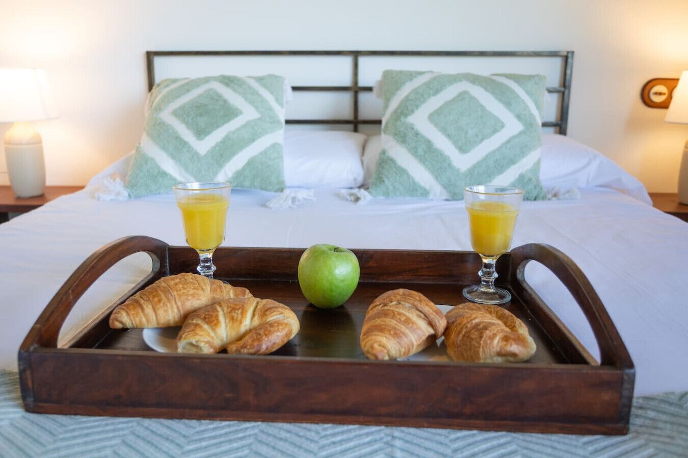 Una cama con almohadas, una bandeja con croissants y una manzana, y vasos de jugo de naranja.