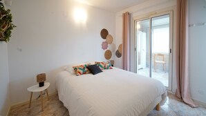 Une chambre avec un grand lit, une petite table de chevet, une lampe fixée au mur et une vue sur l’extérieur grâce à une porte-fenêtre coulissante.