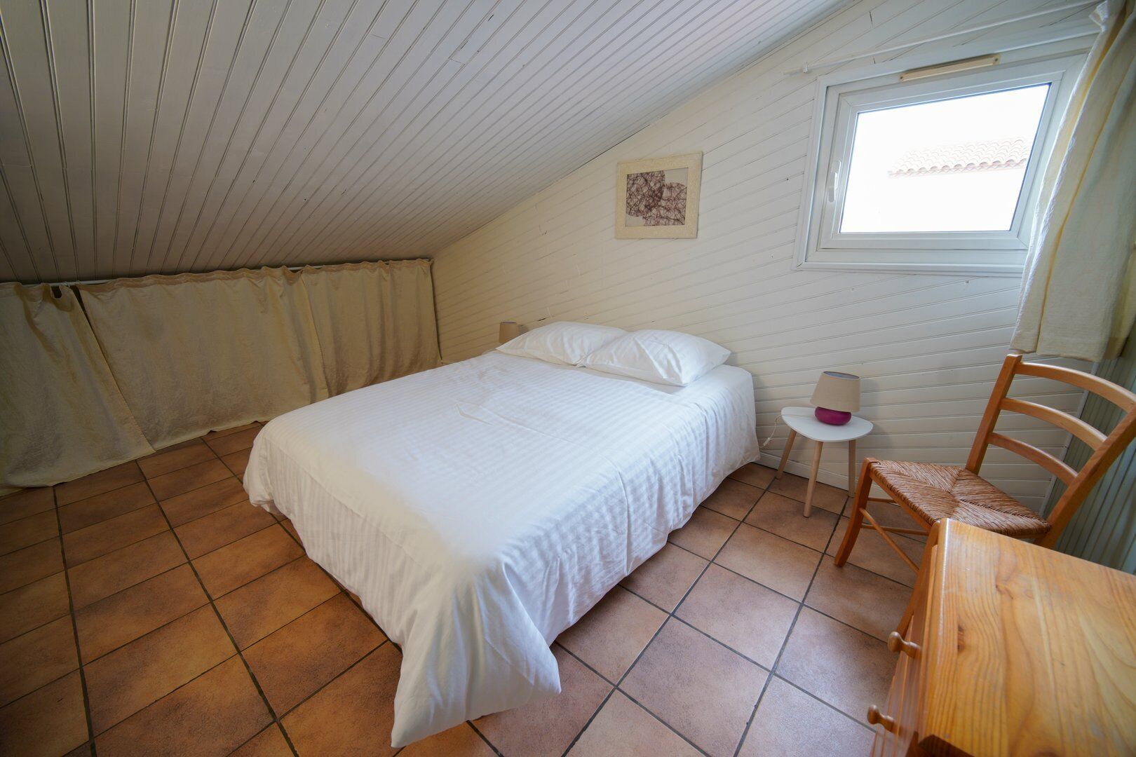 A bedroom with a bed, a chair, a small table, a window, and a picture on the wall.