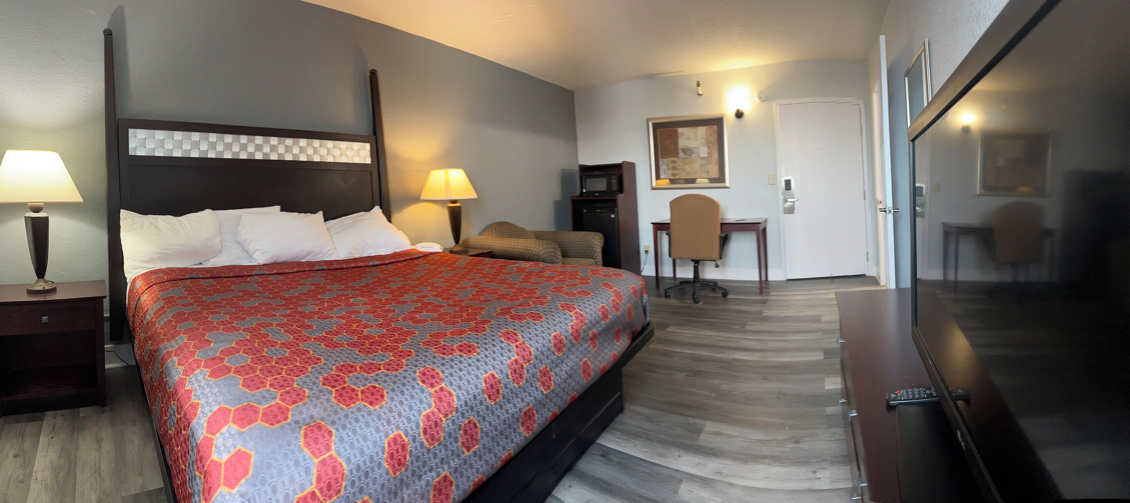 A hotel room with a large bed, a desk, a chair, a TV, and a door leading to another room.