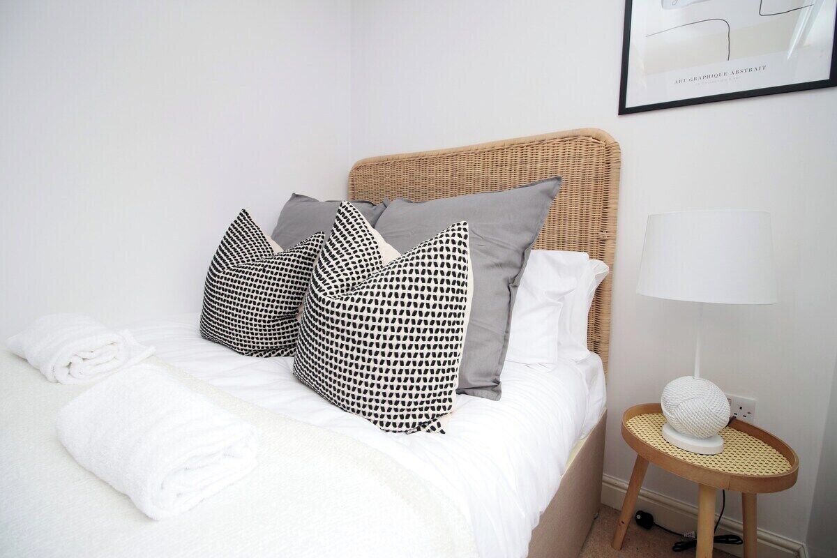 Un lit bien fait, avec du linge de lit blanc, une tête de lit en osier et des coussins à motifs. Une petite table ronde avec une lampe blanche et un objet sphérique se trouve à côté du lit.
