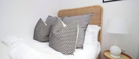 A neatly made bed with white linens, a wicker headboard, and patterned pillows. A small round table with a white lamp and a spherical object is beside the bed.