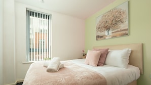A neatly made bed with a pink blanket and pillows, a window with blinds, and a painting of a tree on the wall.