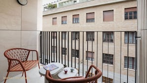 Un balcón con sillas de mimbre, una mesita pequeña y una revista. Al fondo, un edificio con varias ventanas y balcones.