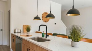 Une cuisine moderne dotée d’un plan de travail en marbre blanc, de meubles en bois, d’un évier en acier inoxydable et de deux luminaires suspendus.