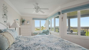 A bedroom with a large bed, a ceiling fan, a window with a view of the beach, and a small table with a lamp. - Ocean Eight 101 by Distinctive Beach Rentals (St. Augustine)