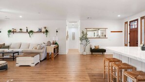 A spacious living area with a sofa, coffee table, and wooden flooring. There are plants on shelves and a dining area with stools. - Escondido Oasis | Sleeps 10 + Hot Tub! (Escondido)