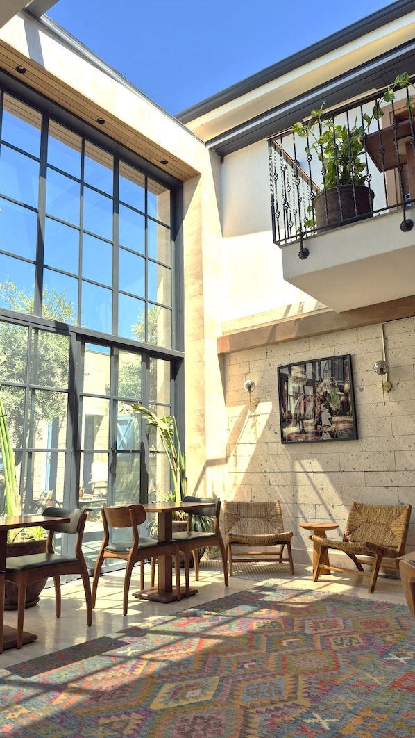 Un espacio interior amplio con ventanas grandes, una alfombra colorida, muebles de madera y un balcón con plantas.