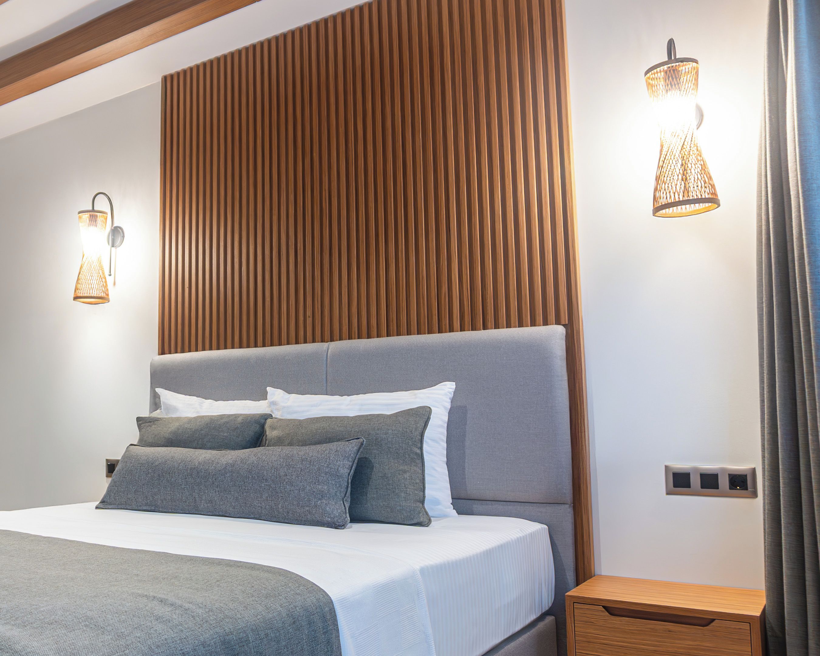 A modern bedroom with a grey upholstered bed, a wooden headboard, and two wall-mounted light fixtures.