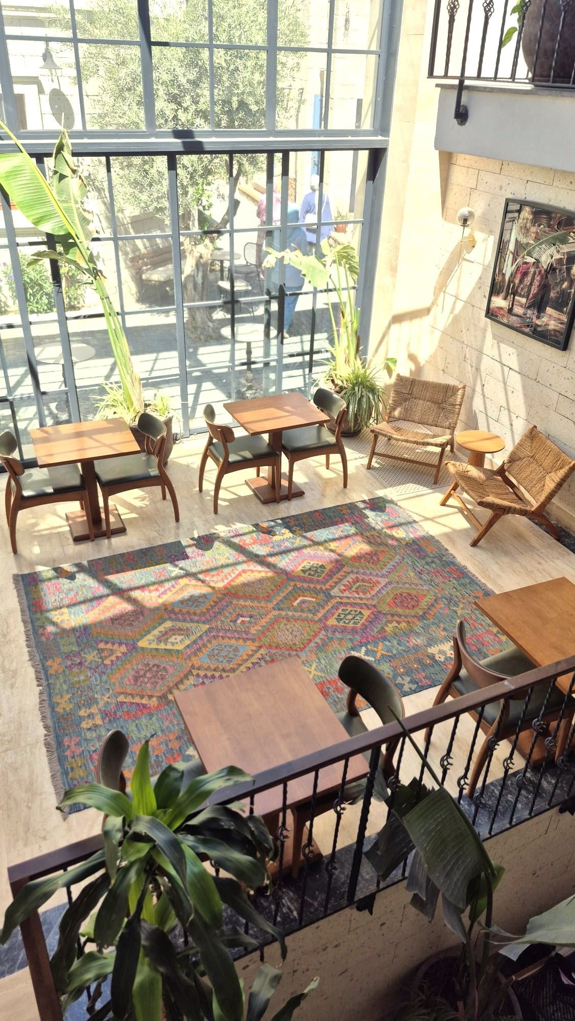 A sunlit indoor space with a wooden table and chairs, a colorful rug, and a balcony with a railing.