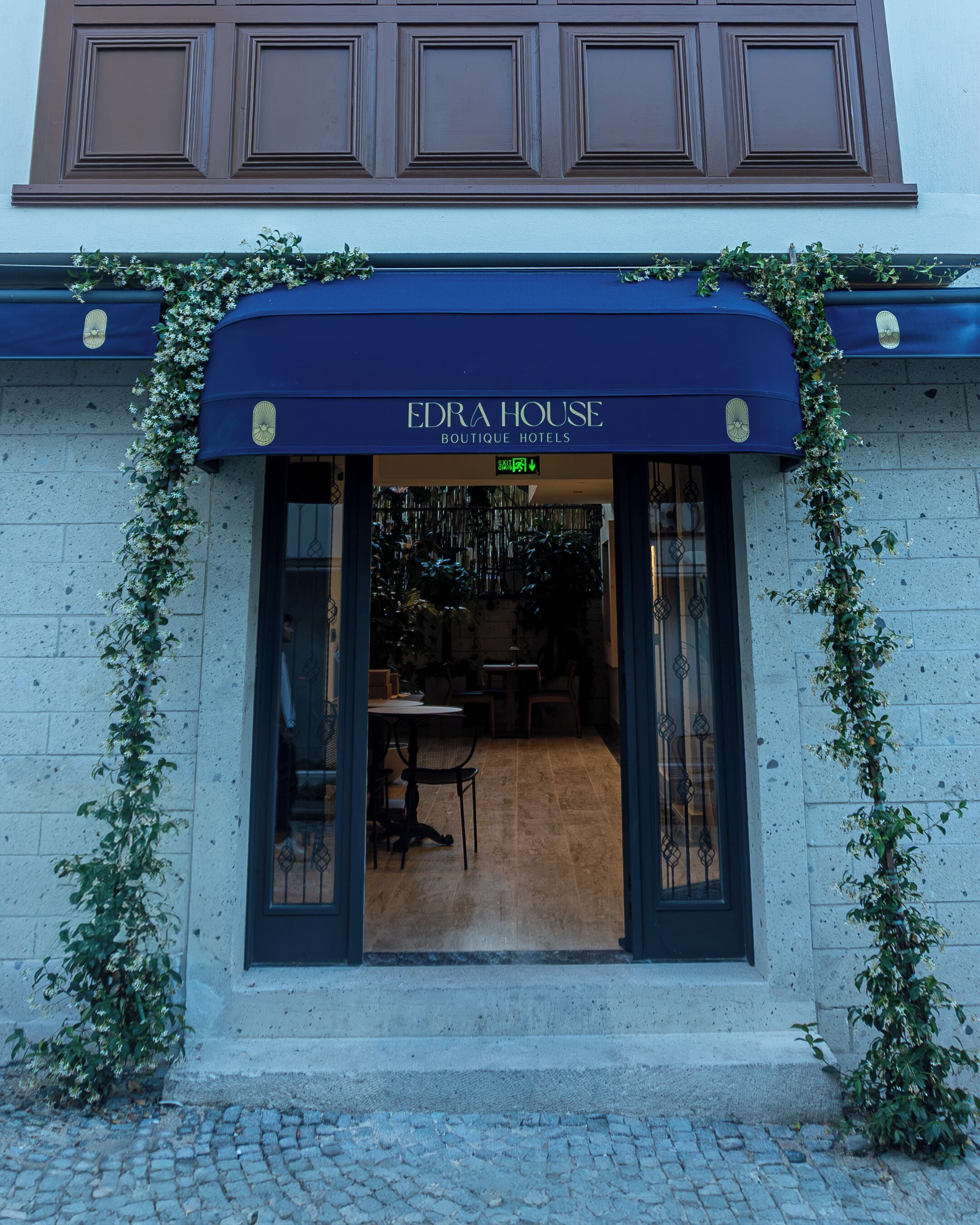 A boutique hotel entrance with a dark blue awning, a sign reading 'EDRA HOUSE BOUTIQUE HOTELS,' and a view into the interior.