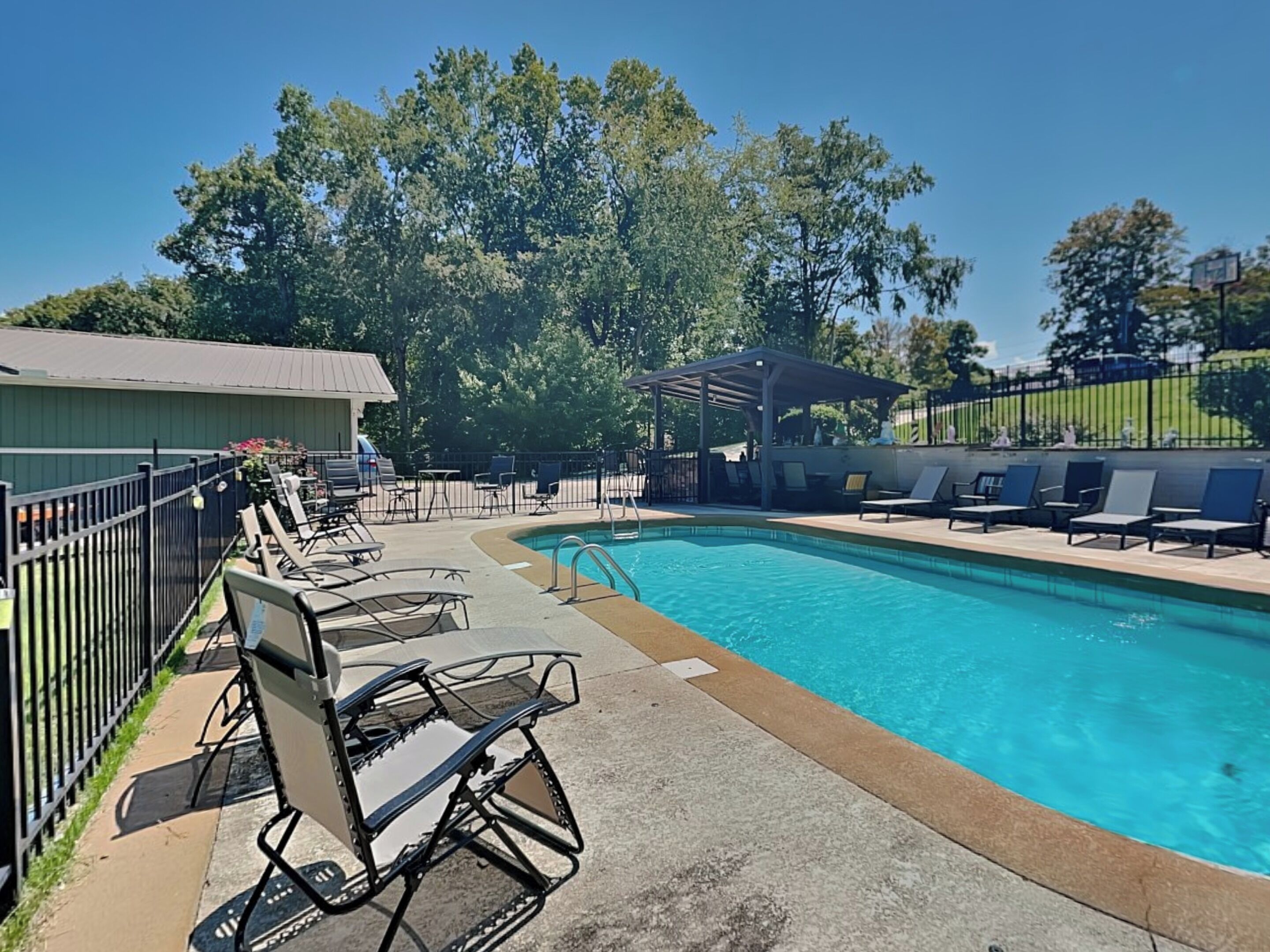 A swimming pool with lounge chairs and a covered seating area, surrounded by trees and a fenced patio.
