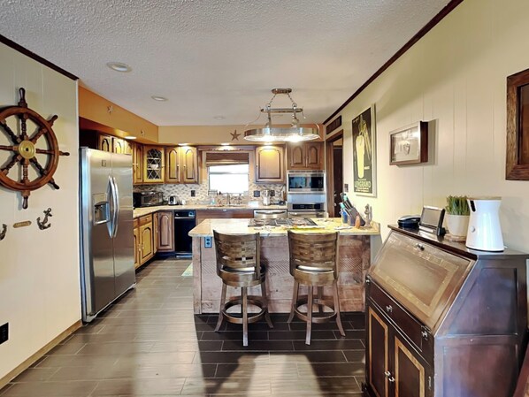 A modern kitchen with a central island, stainless steel appliances, wooden cabinets, and a ship's wheel decoration. - Sunset Cove by Jackson Mountain Rentals (Dandridge)