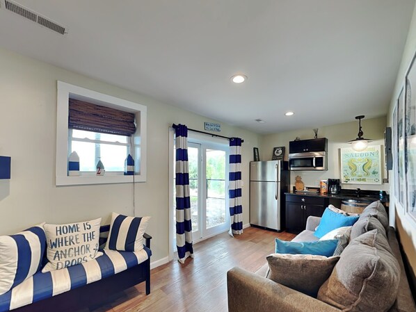 A modern living room with a sofa and armchair, a kitchen with a refrigerator and microwave, and a window with blinds.