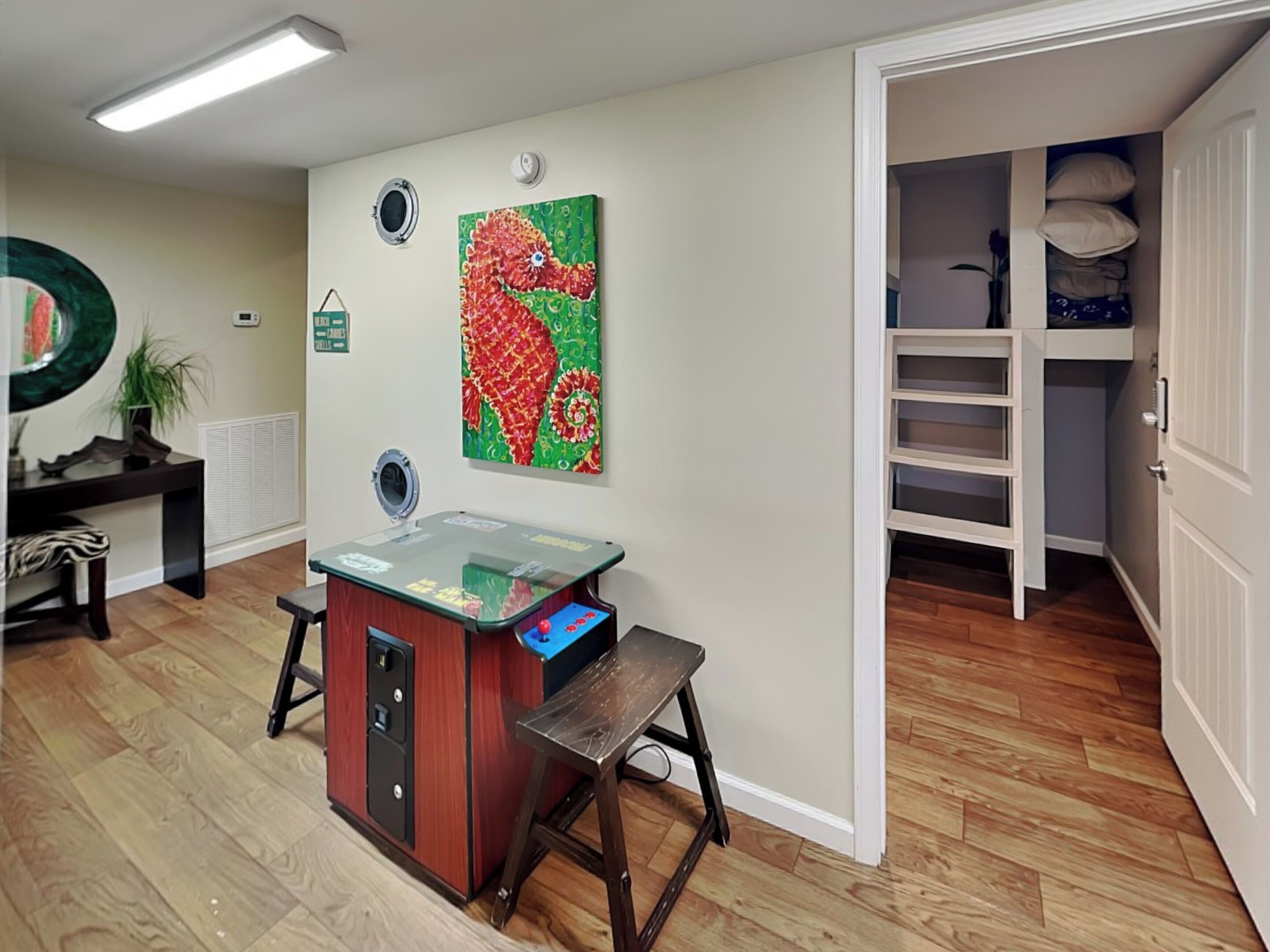 A room with a wooden floor, a desk with a computer, a chair, a small table, and a wall with a colorful painting.
