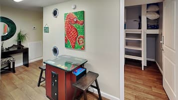 A room with a wooden floor, a desk with a computer, a chair, a small table, and a wall with a colorful painting.