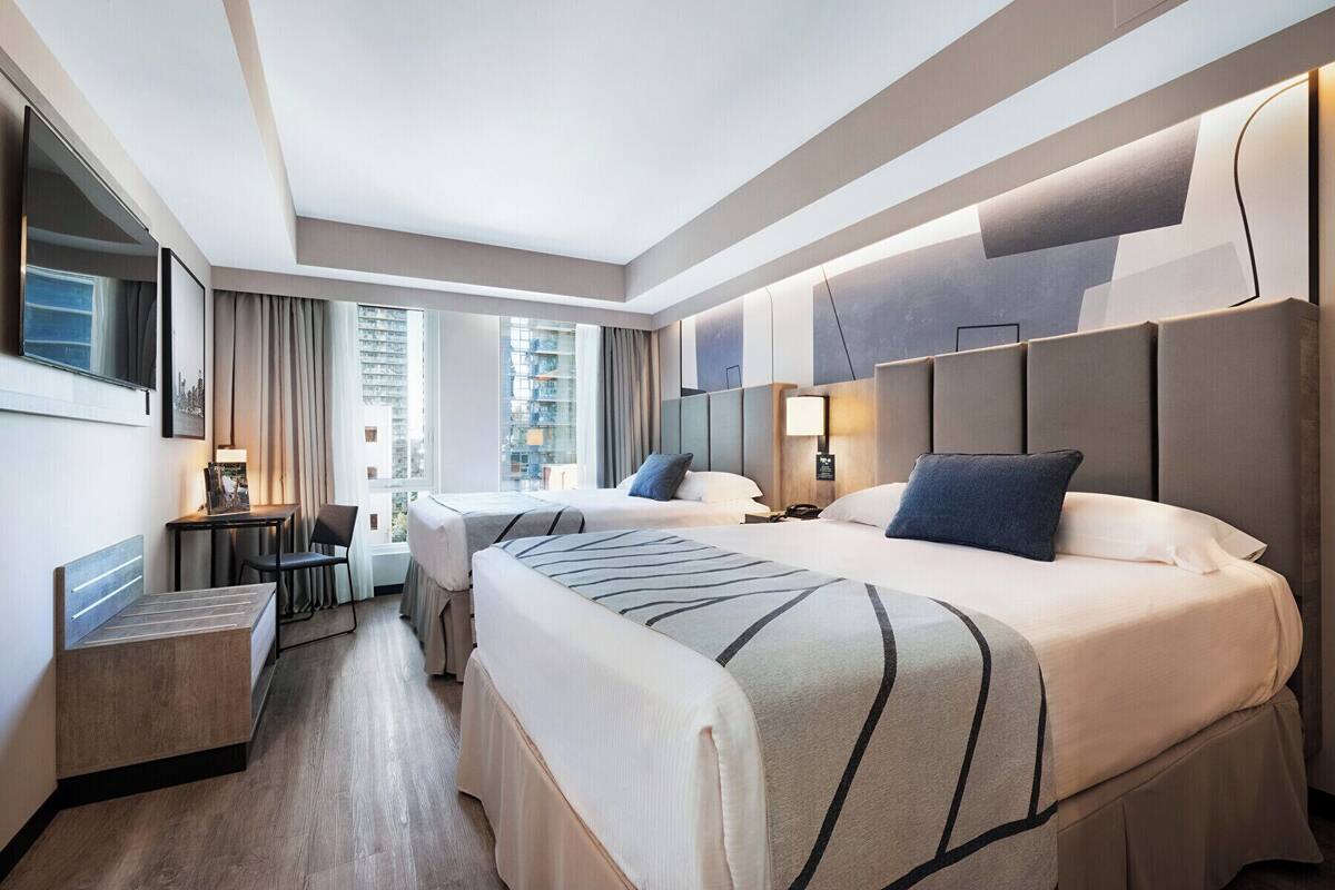 A modern hotel room with two beds, a desk, and a large window offering a city view.