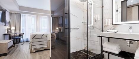 A modern hotel room with a glass-enclosed shower, a marble-tiled bathroom, a desk with a chair, and a bed with a striped blanket.