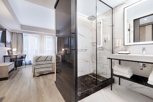Een moderne hotelkamer met een douche in een glazen hoek, een betegelde badkamer met marmer, een bureau met stoel en een bed met een gestreept dekbed.