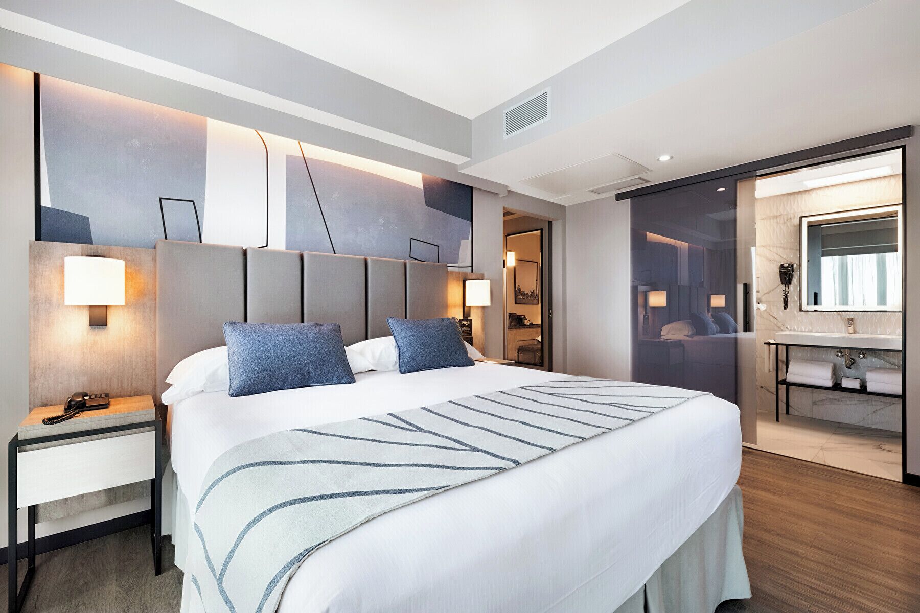A modern hotel room with a large bed, bedside tables, a built-in desk, a large mirror, and a bathroom with a vanity and sink.