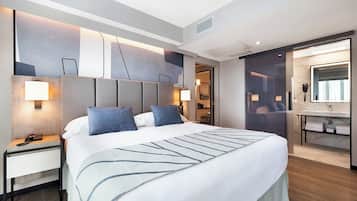 Et moderne hotelværelse med en stor seng, sengeborde, et indbygget skrivebord, et stort spejl og et badeværelse med vaskebord og håndvask.