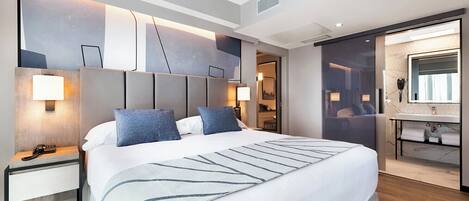 A modern hotel room with a large bed, bedside tables, a built-in desk, a large mirror, and a bathroom with a vanity and sink.