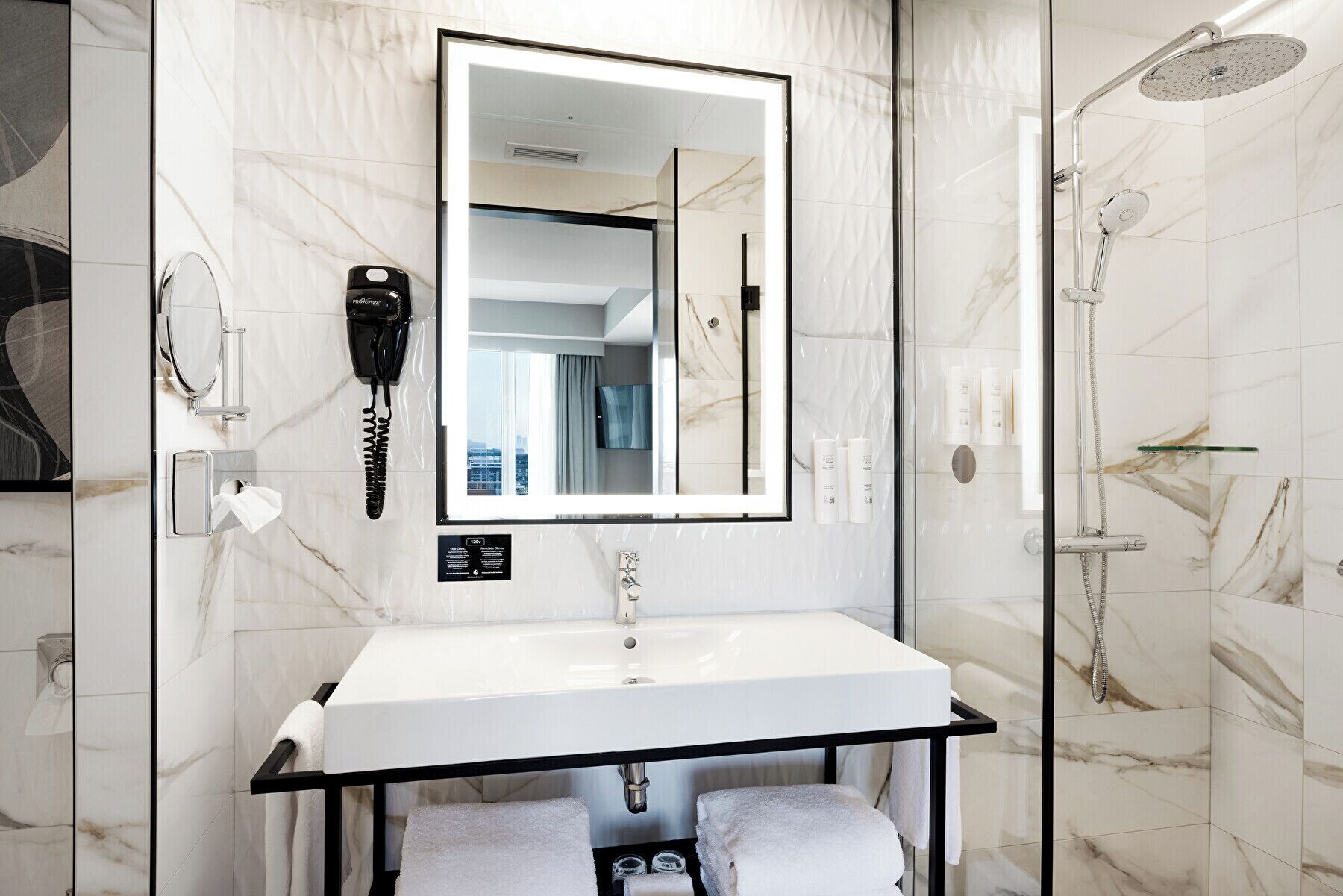 A modern bathroom with a marble backsplash, a large mirror, a white sink, a handheld showerhead, and a hairdryer mounted on the wall.