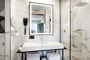 Een moderne badkamer met een marmeren spatwand, een grote spiegel, een wit wastafel en een handdouche. Een haardroger is aan de muur bevestigd.