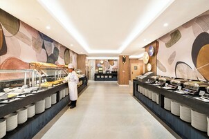 Uma área de buffet com um chef, um balcão com diversos alimentos e uma receção ao fundo.