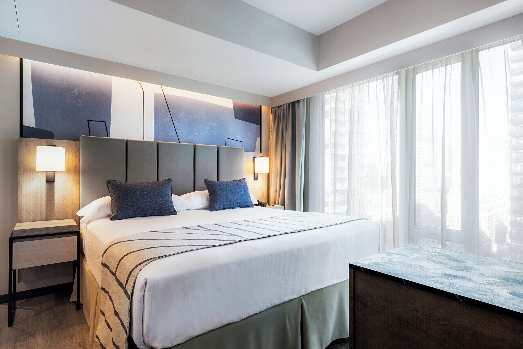 A modern hotel room with a large bed, two bedside lamps, a desk, and a view of the city through large windows.