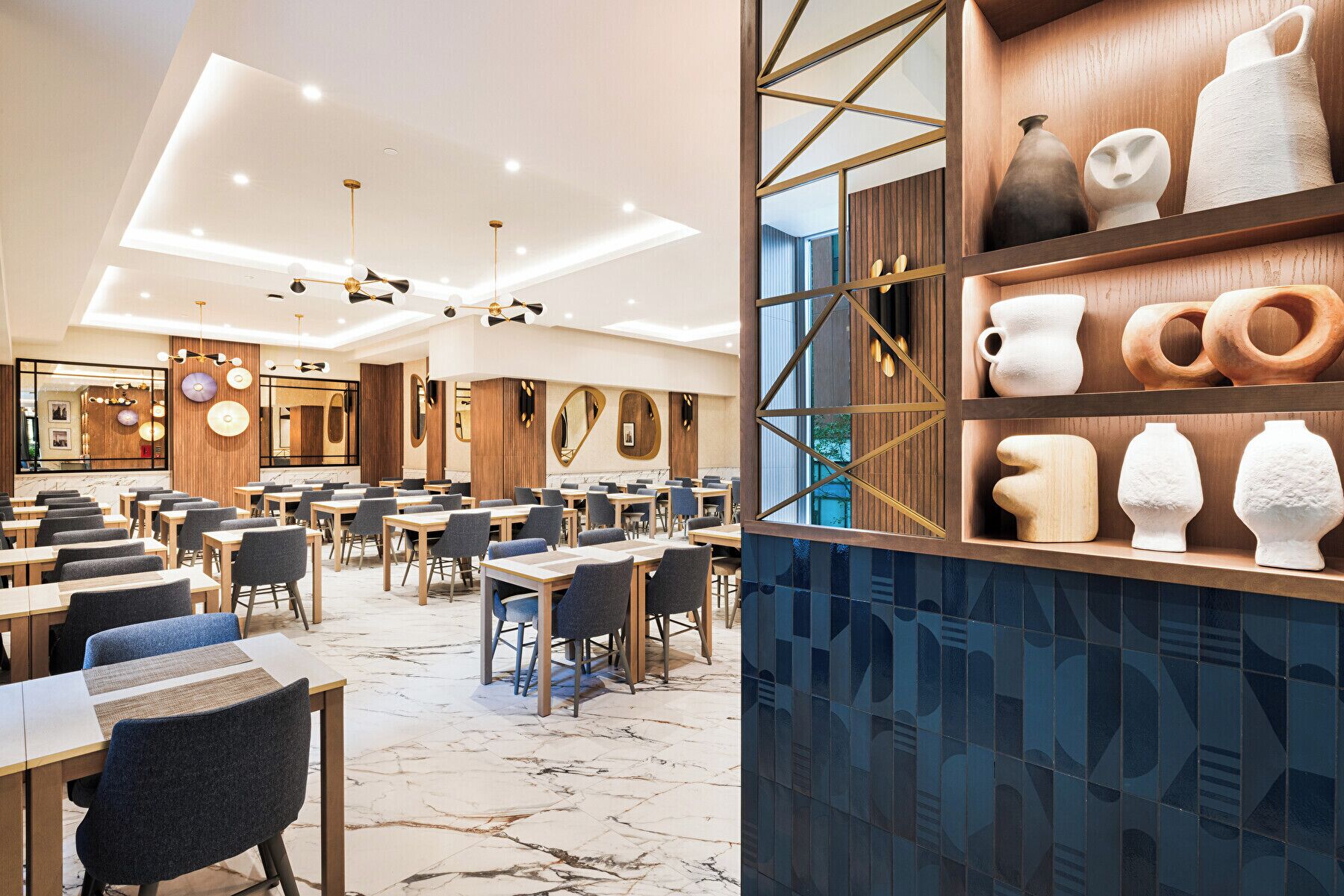 A modern dining area with multiple tables and chairs, a central chandelier, and a wall with shelves displaying various ceramic items.