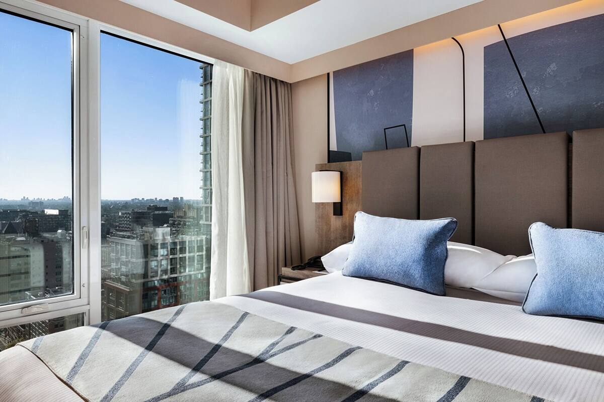 A hotel room with a large bed, a nightstand, a lamp, and a view of the city through large windows.