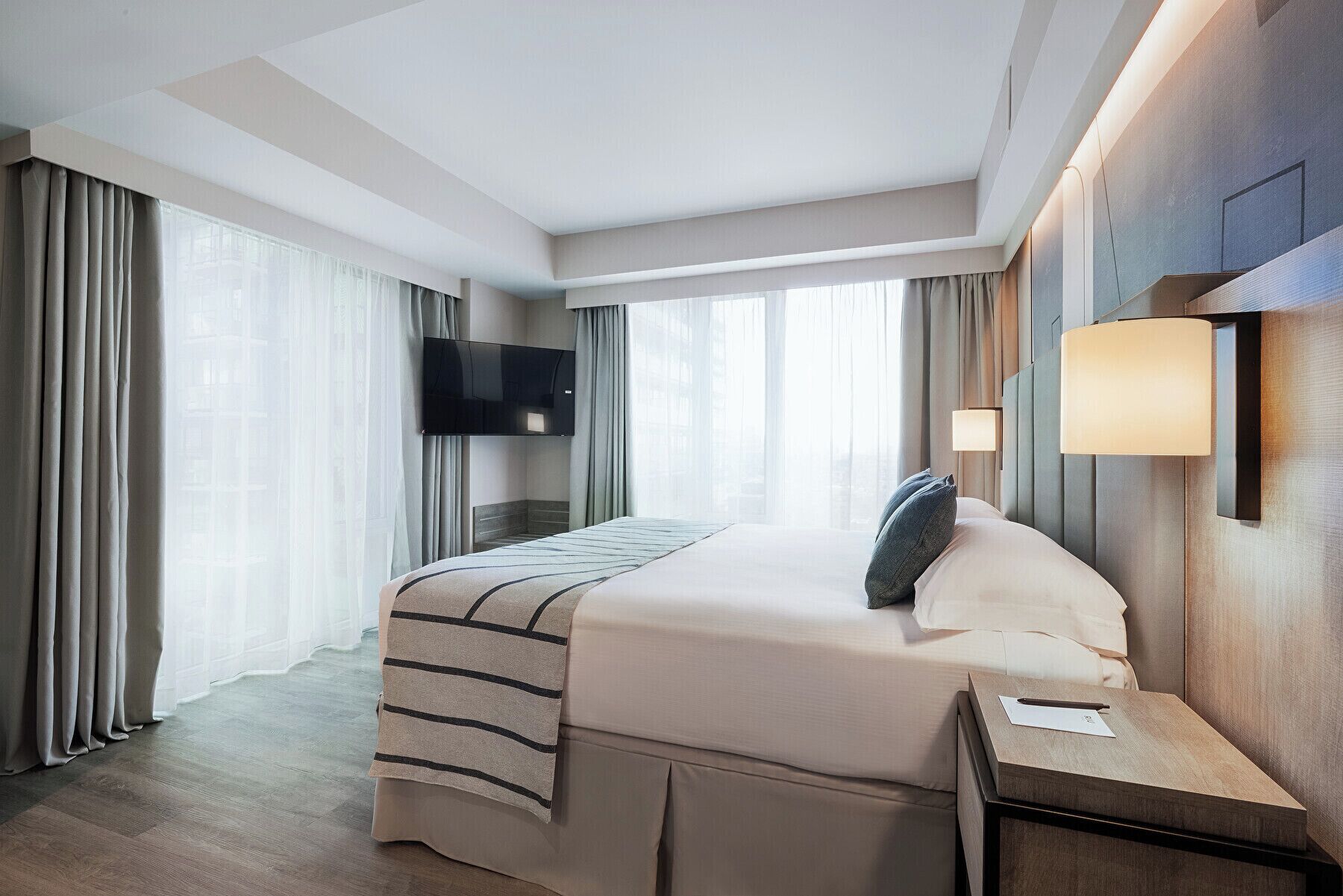 A hotel room with a large bed, a flat-screen TV mounted on the wall, and a bedside table with a lamp.