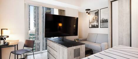 A hotel room with a bed, a desk with a flat-screen TV, a sofa, a chair, and a large window offering a city view.