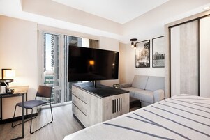 Een hotelkamer met een bed, een bureau met een flatscreen televisie, een bank, een stoel en een groot raam met uitzicht over de stad.