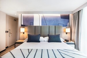 Een hotelkamer met een groot bed, twee nachtkastjes met lampen, een hoofdbord met abstracte kunst en uitzicht op de stad door het raam.