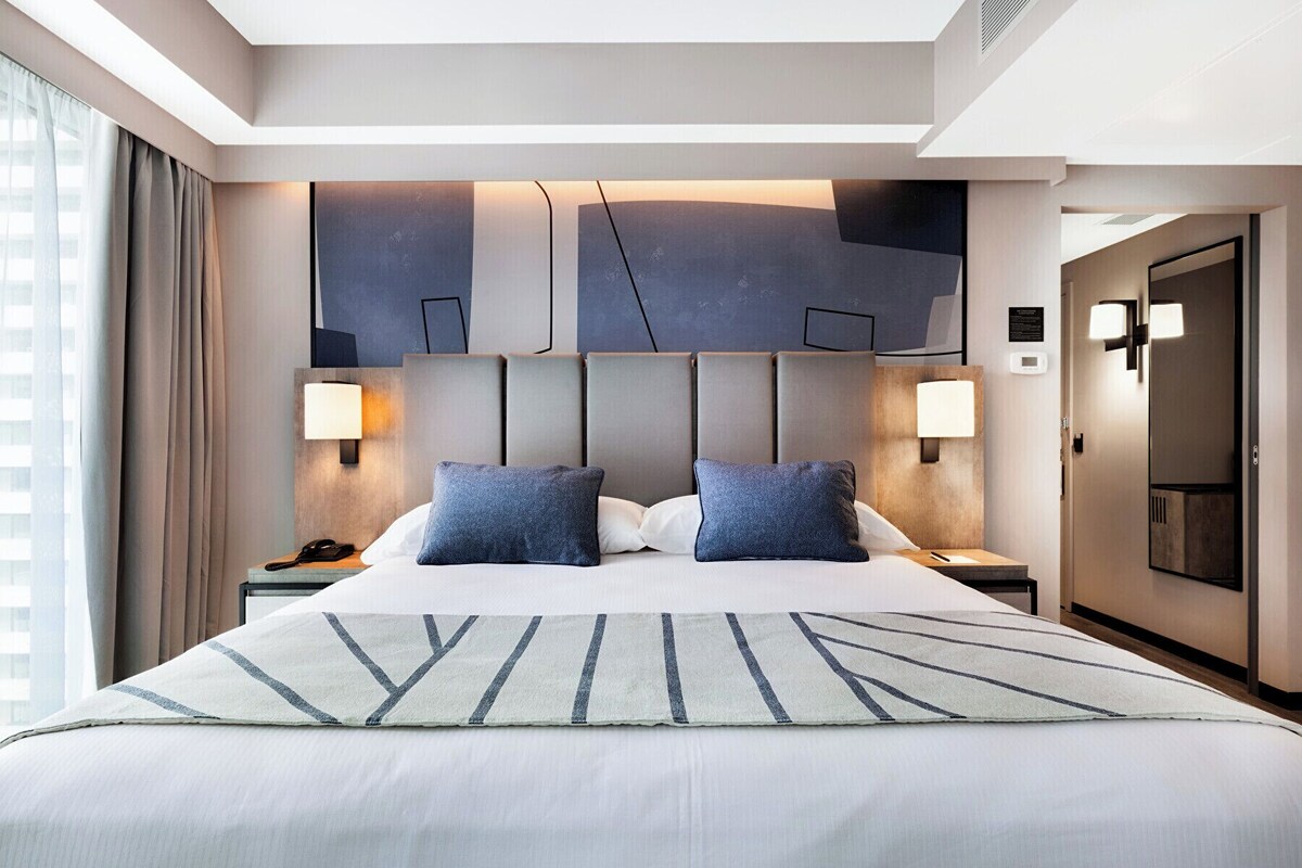 Ein modernes Hotelzimmer mit einem großen Bett, zwei Nachttischlampen, einem Kopfteil mit geometrischen Mustern und Blick auf die Stadt durch das Fenster.