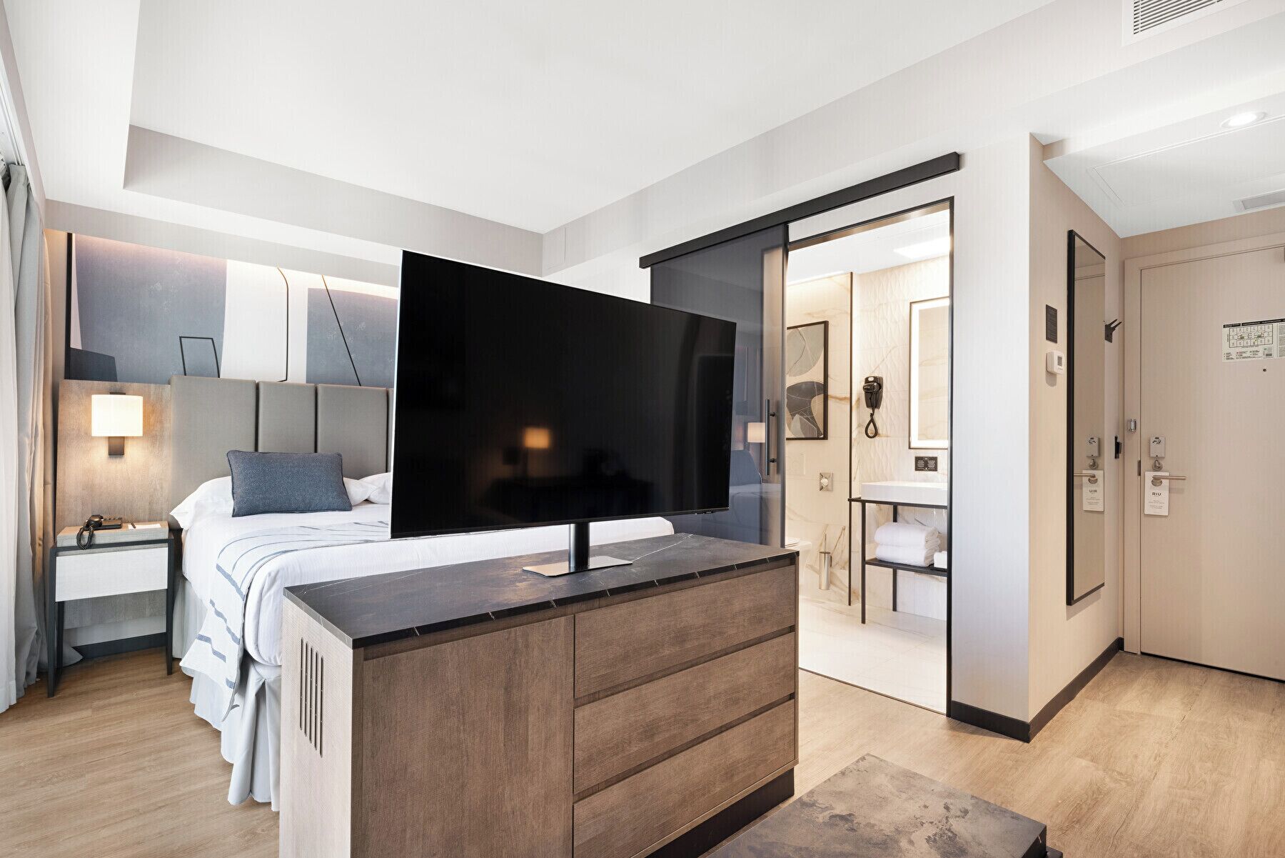 Una habitación de hotel moderna con un televisor de pantalla plana grande, un aparador de madera, una cama con ropa blanca y un baño visible a través de una puerta de vidrio.