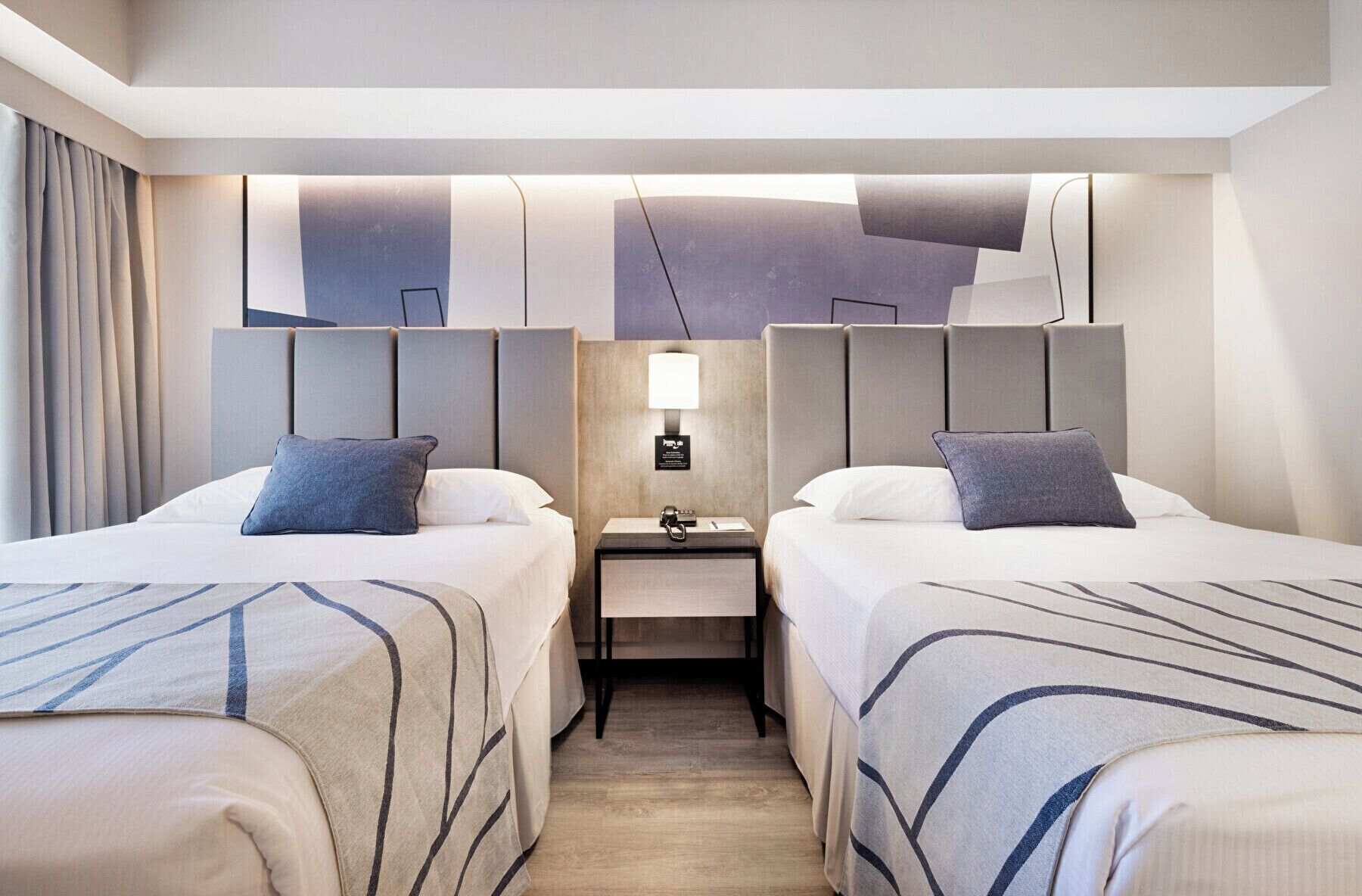 A hotel room with two beds, a nightstand, a lamp, and a wall-mounted light fixture.