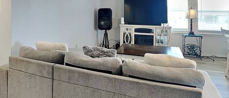 A modern living room with a grey sectional sofa, a flat-screen TV, a speaker, and a small side table with a lamp.